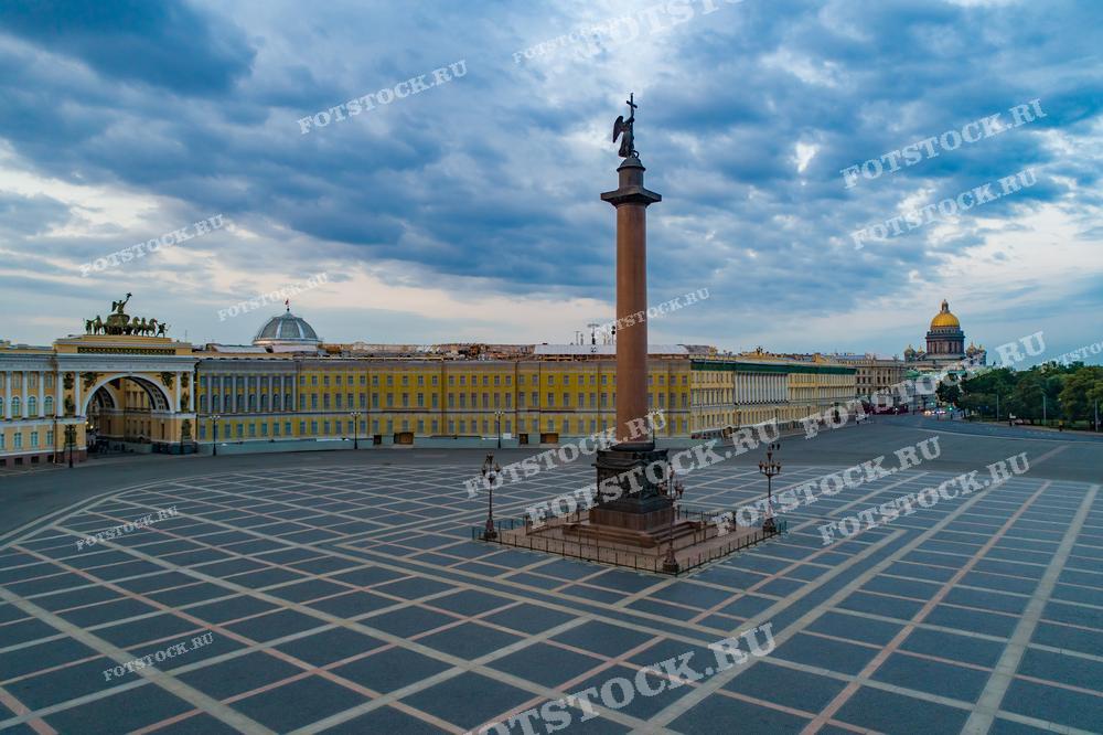 На фотографии изображена Дворцовая площадь в Санкт-Петербурге под пасмурным небом. Основные элементы снимка — Александровская колонна в центре, Главный штаб с аркой и купол Исаакиевского собора. Это изображение подойдет для использования в образовательных и исторических материалах, туристических брошюрах, рекламных кампаниях, а также в статьях о городском планировании и архитектуре.